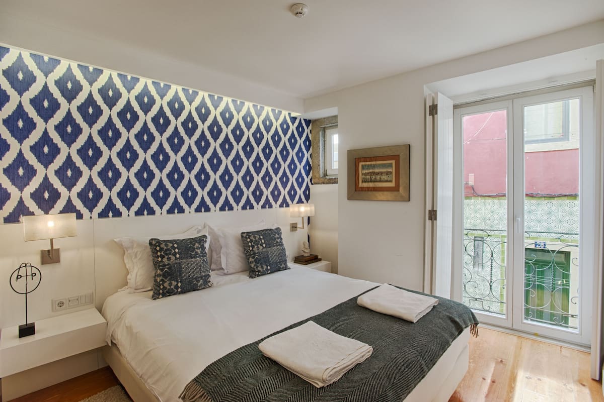 Un dormitorio con pared estampada, una cama con dos almohadas, una mesita de noche con lámpara y una ventana con vista a un edificio rojo.