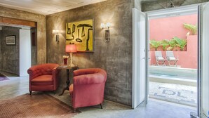 Un vestíbulo con un sillón rojo, una mesita con una lámpara, un cuadro y vistas del patio exterior con sillas.