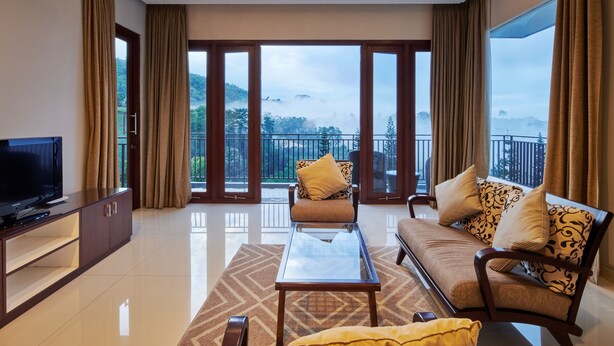 Una sala de estar moderna con televisión, sofá y mesa de centro, ventanas amplias con cortinas y un balcón con vistas a las montañas.