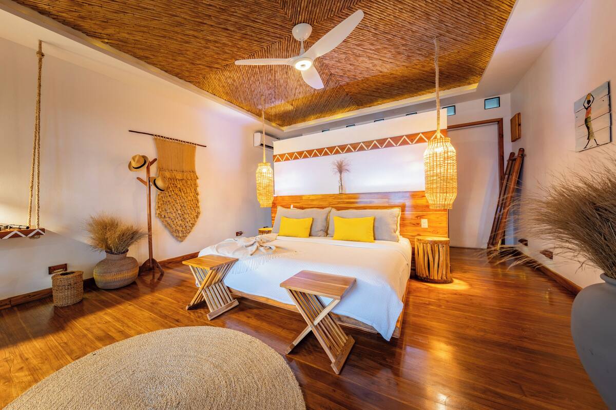 Een slaapkamer met een houten plafond, een bed met gele kussens, een hanglamp en een grote vaas met gedroogde planten.