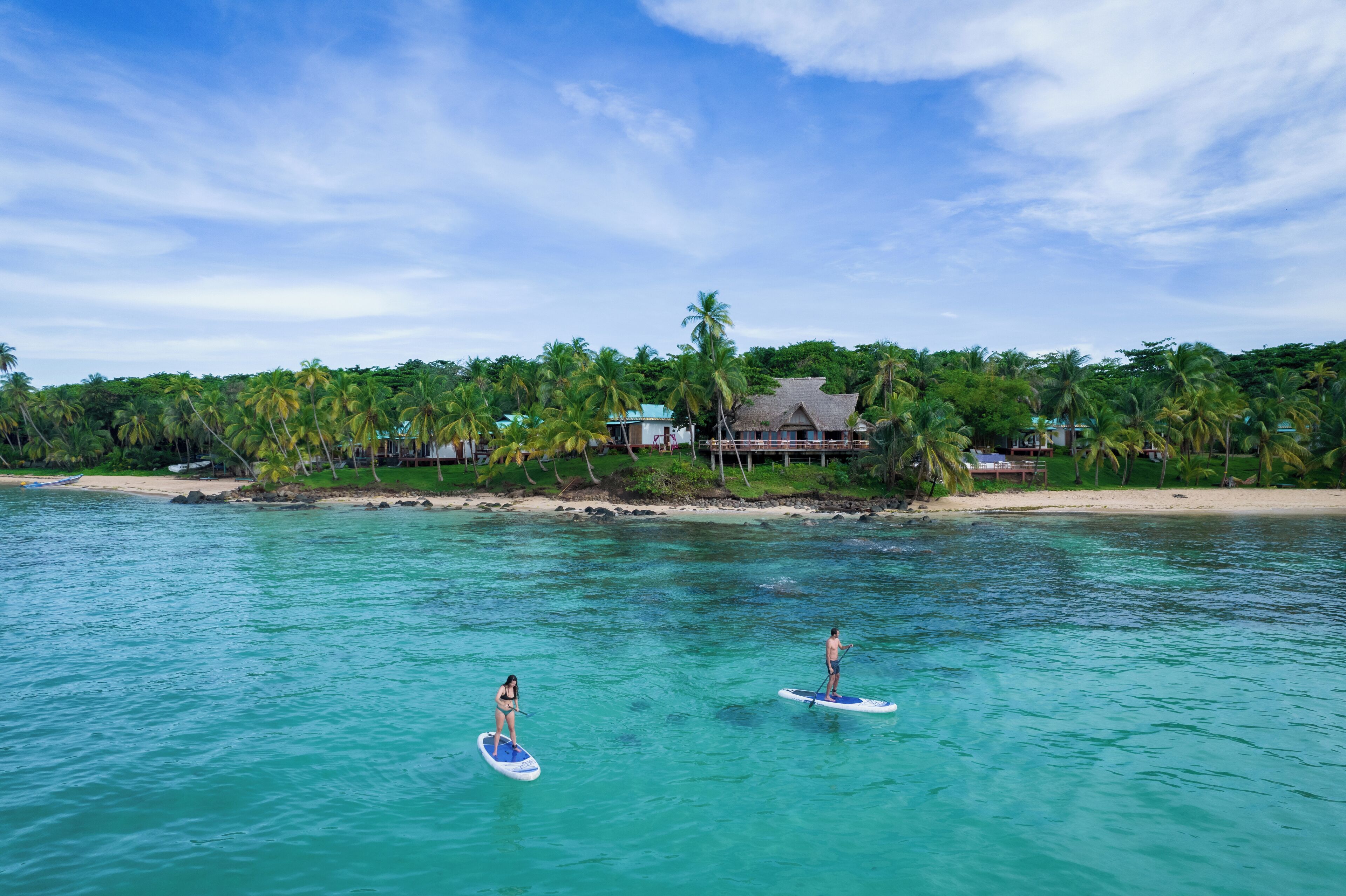 A beach with clear turquoise water, two people paddleboarding, and a house with a thatched roof on the shore.
