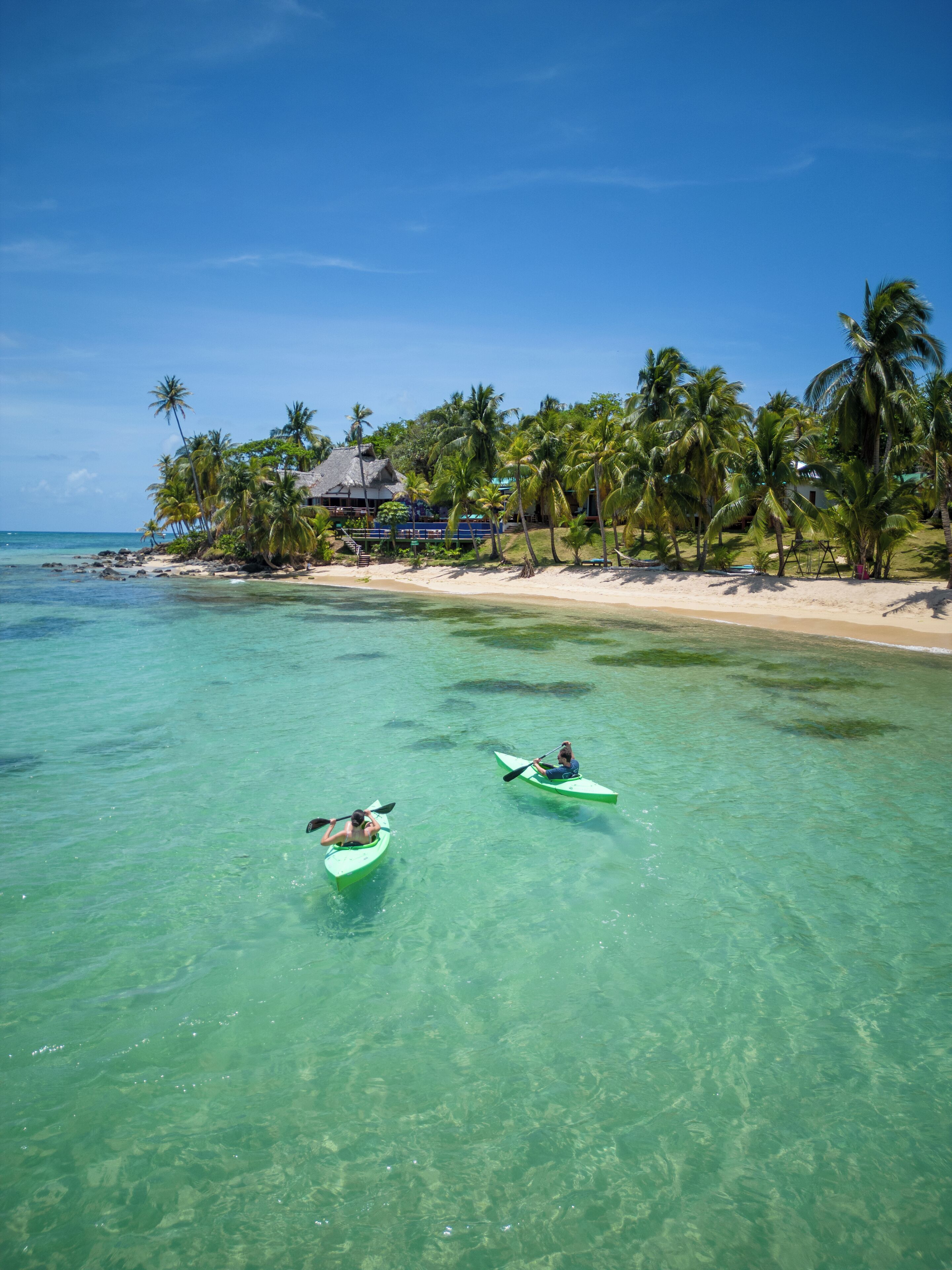 Two kayaks on clear turquoise water with a beach and palm trees in the background.