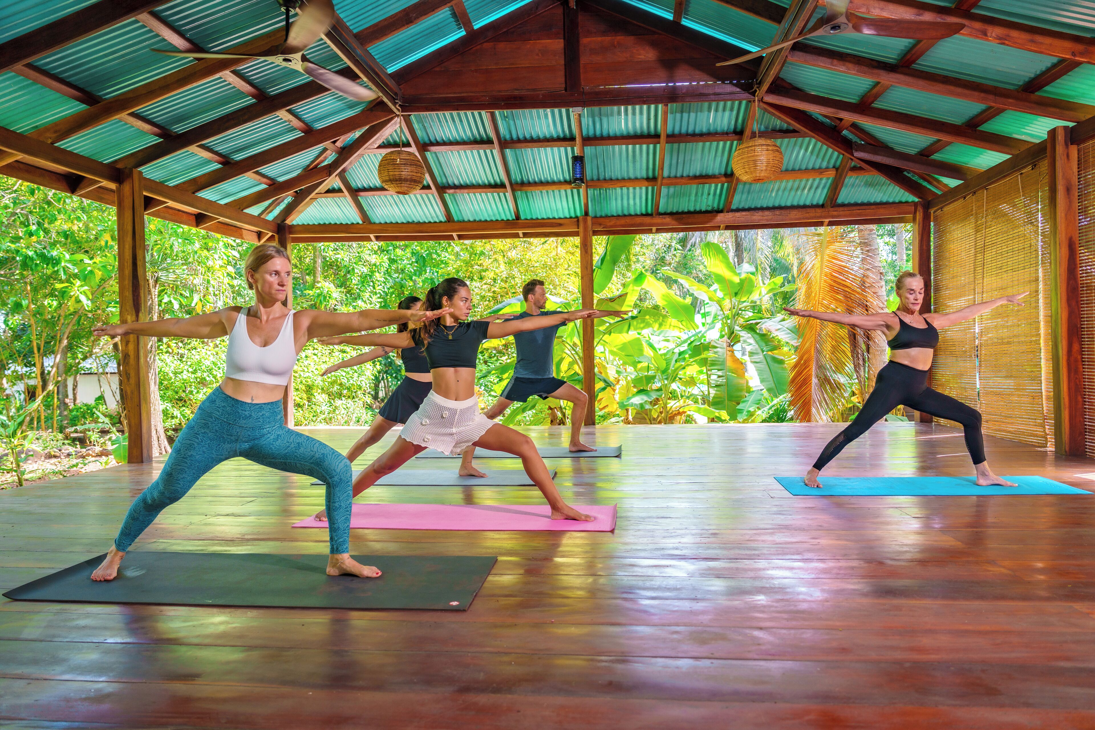 A group of people practicing yoga in a covered area with a wooden floor and a metal roof.