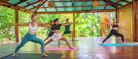 A group of people practicing yoga in a covered area with a wooden floor and a metal roof.