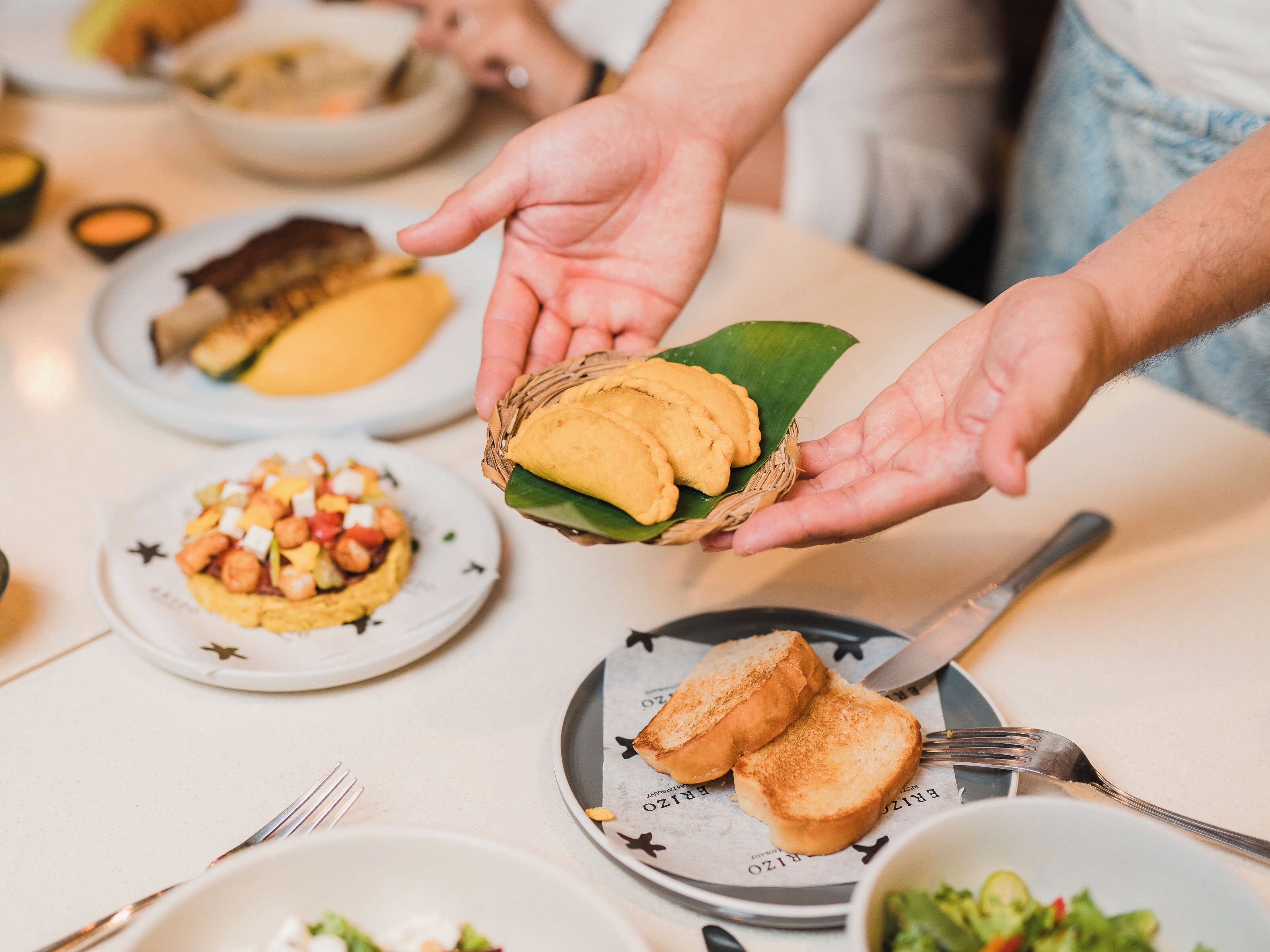 A person holding a basket with a sandwich and a leaf, with various dishes on a table.