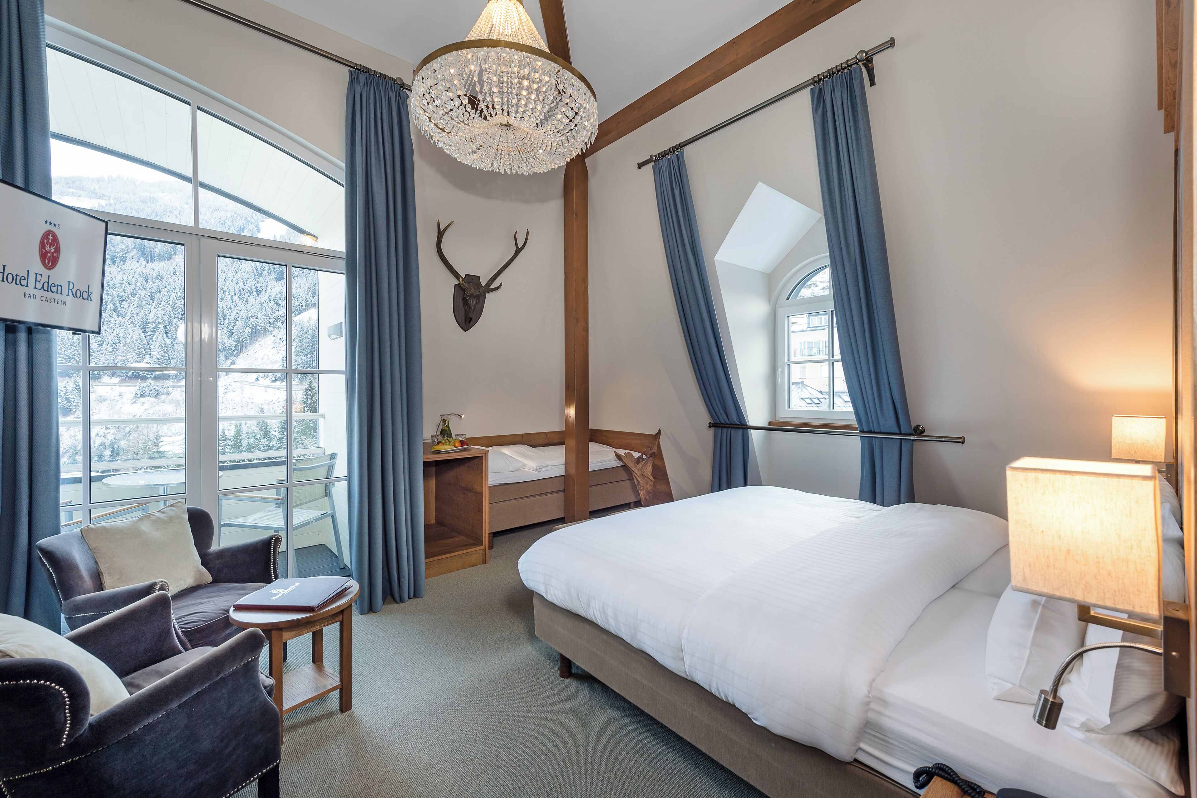 A hotel room with a bed, a chair, a desk, a chandelier, and a view of the outdoors.