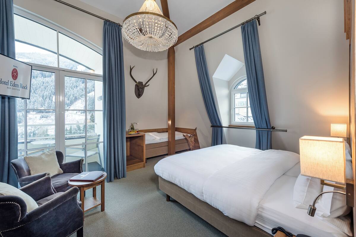 A hotel room with a bed, a chair, a desk, a chandelier, and a view of the outdoors.