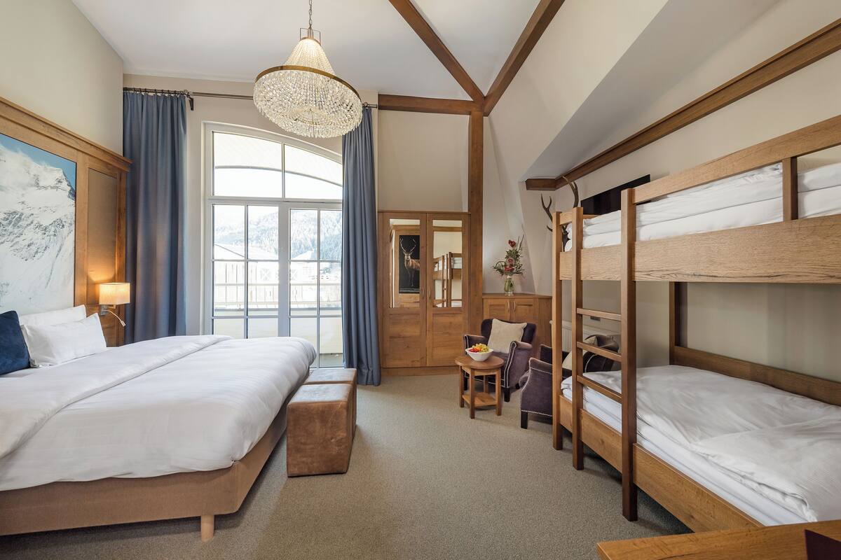 A hotel room with a large bed, a smaller bed, a desk, a chair, a chandelier, and a view of the outdoors.