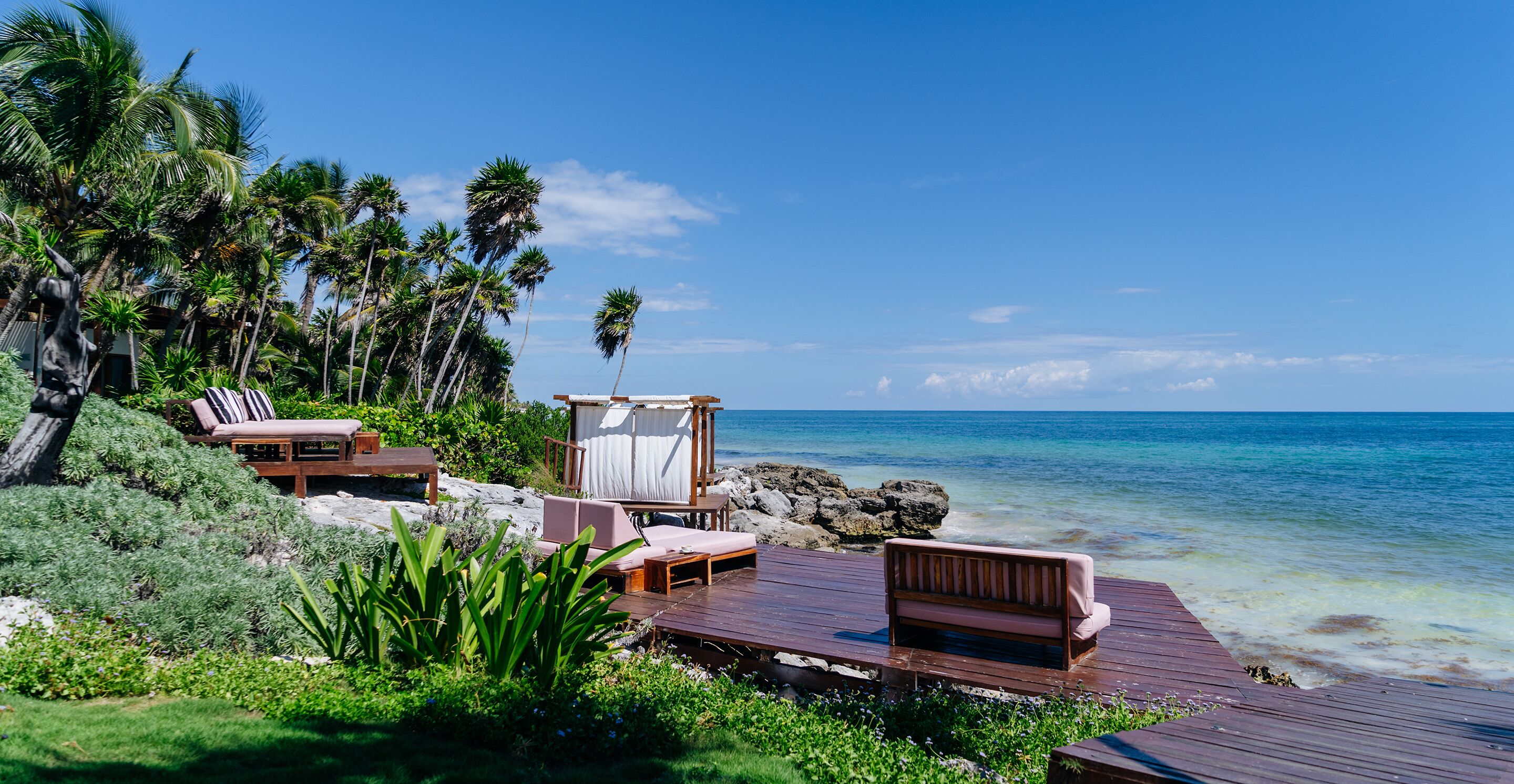A wooden deck with lounge chairs and a covered seating area overlooking a beach with palm trees and clear blue water.