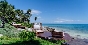 Een houten terras met lounge fauteuils en een overdekte zithoek, met uitzicht op een strand met palmbomen en helderblauw water.