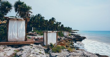 A beachside resort with wooden deck seating, white curtains, and palm trees.