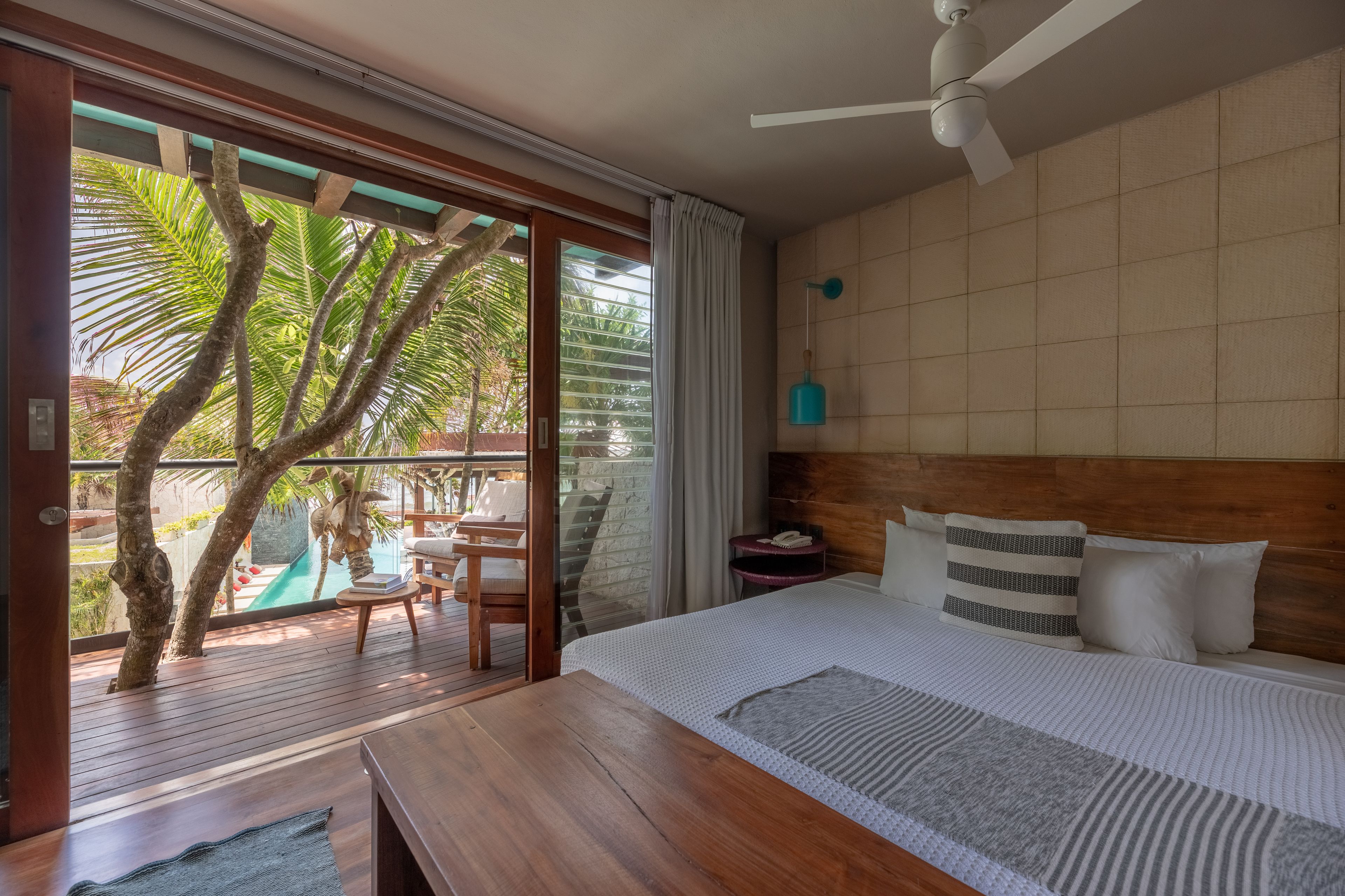 A bedroom with a wooden headboard, a bed with striped bedding, a nightstand, and a view of a pool and palm trees through a sliding glass door.