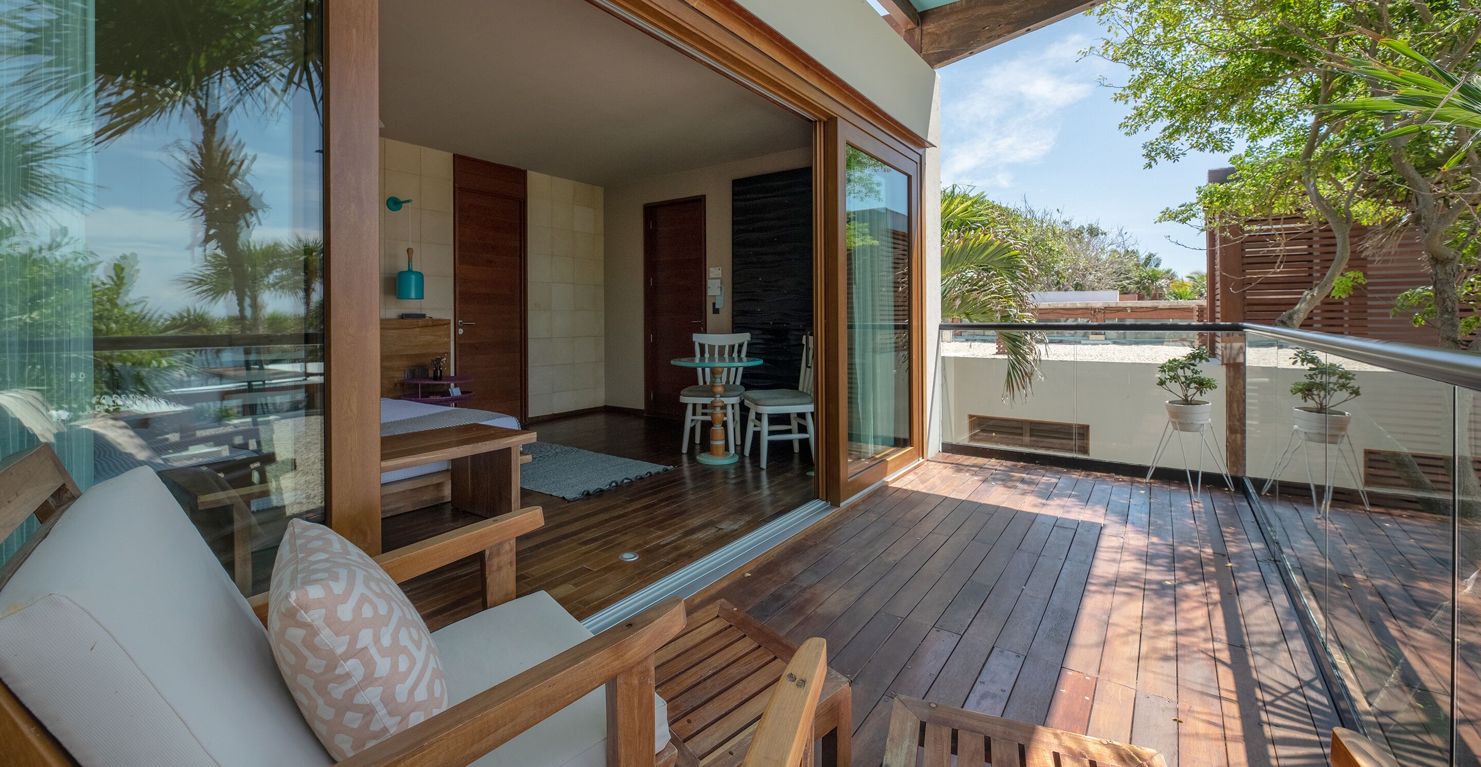 A spacious balcony with wooden flooring, a seating area with cushions, and a glass door leading to a room with a bed and a small table.