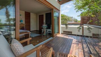 A spacious balcony with wooden flooring, a seating area with cushions, and a glass door leading to a room with a bed and a small table.
