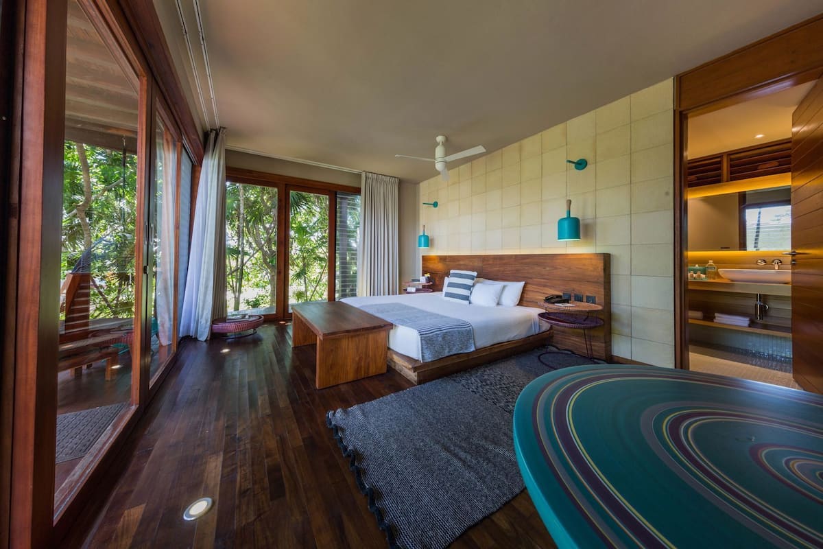Ein modernes Schlafzimmer mit einem großen Bett, einer hölzernen Bank, einem runden Tisch und Blick ins Freie.