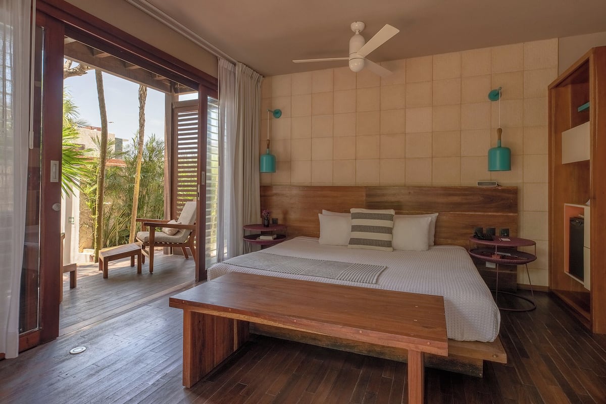 Ein Schlafzimmer mit einem hölzernen Bett, einer Holzbank, einem Deckenventilator und Blick auf Palmen durch die offene Tür.