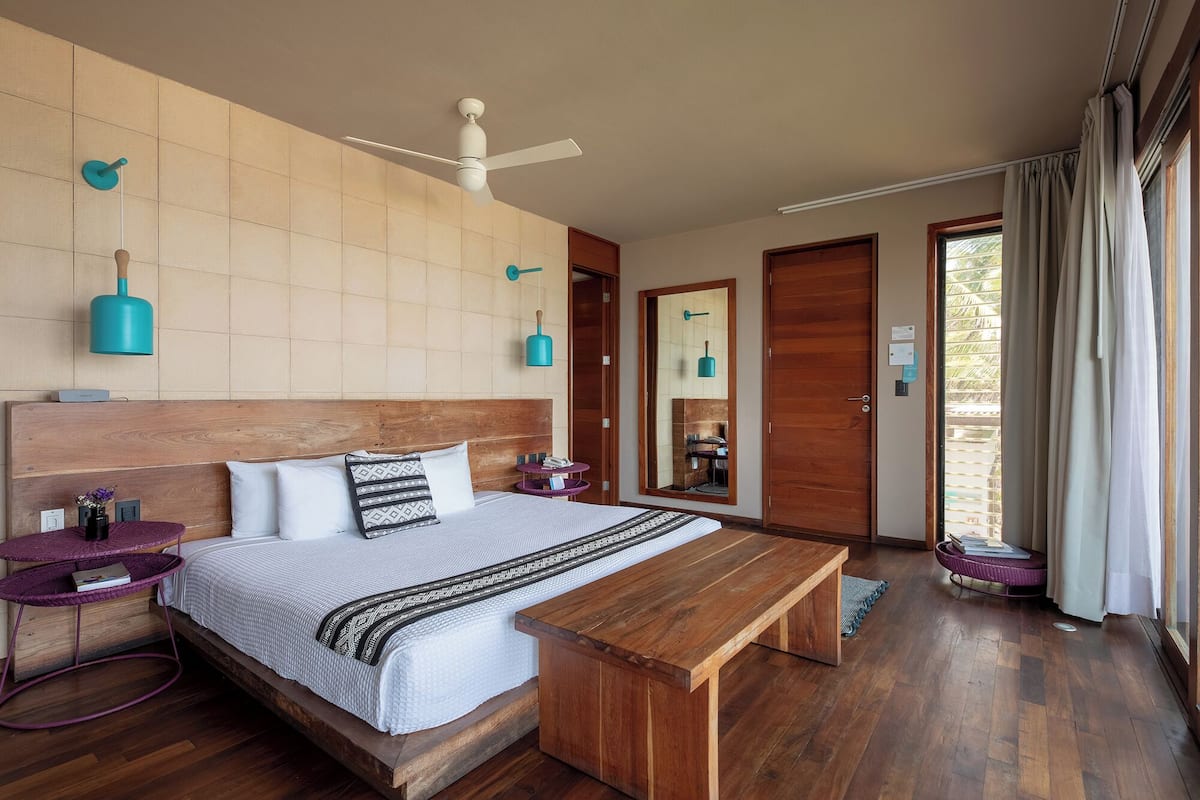 Ein Schlafzimmer mit Holz-Kopfteil, einem Bett mit gemusterten Betttextilien, einer Holzbank, einem Deckenventilator und Blick ins Freie durch eine Tür.