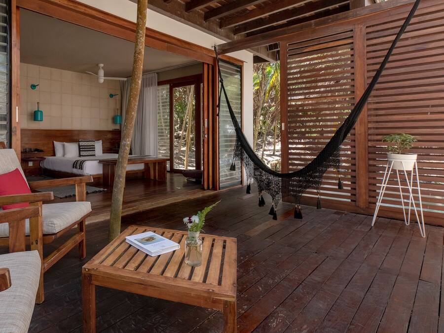 Una terraza de madera con una hamaca, una mesa de madera con un libro y un cojín rojo en una silla.