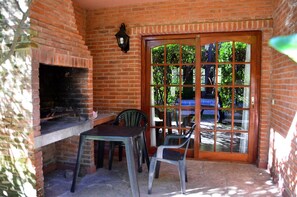 A brick patio with a grill, two plastic chairs, and a wooden table. - Vacances Dorado (Pinamar)