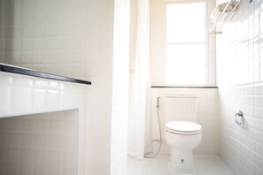 Une petite salle de bain blanche, avec des toilettes, un espace douche et une fenêtre.