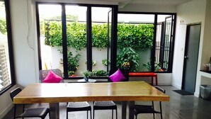 A dining area with a wooden table and chairs, a bench, and a view of a green garden through large windows. - Vanilla Residence Chiangmai (Chiang Mai)