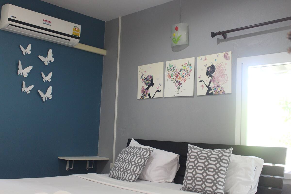 A bedroom with a bed, patterned pillows, wall-mounted air conditioning, butterfly decorations, and floral artwork.