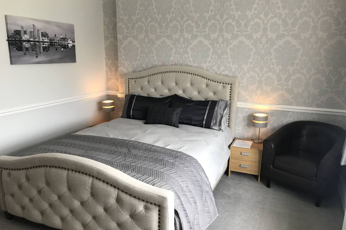A neatly arranged bedroom with a tufted headboard, a cityscape artwork on the wall, and two bedside lamps.
