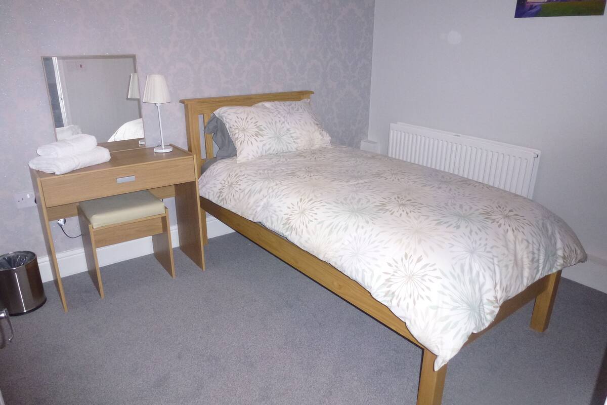 A bedroom with a single bed, a wooden desk with a chair, a mirror, and a radiator.