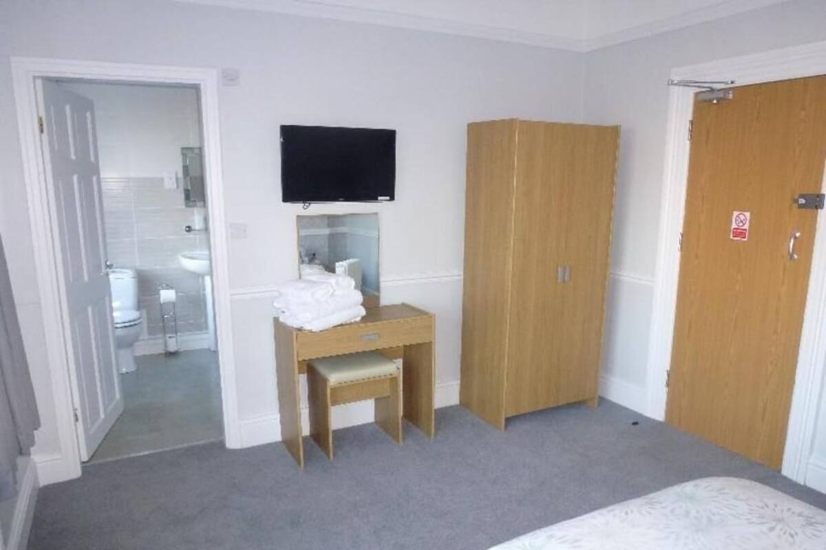A bedroom with a bed, a wooden wardrobe, a desk with a chair, a television mounted on the wall, and a door with a 'No Entry' sign.