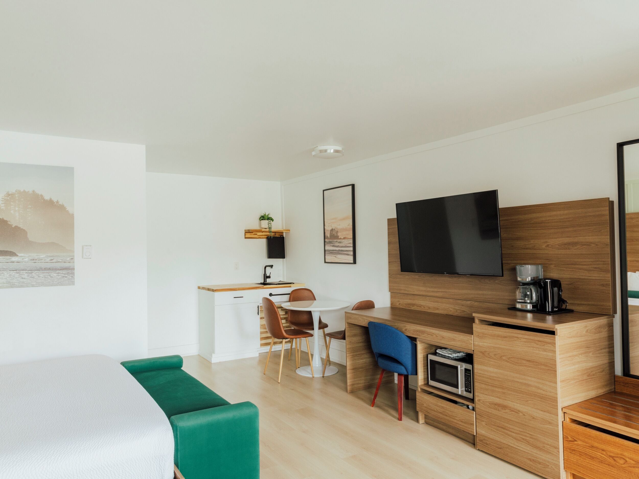 A modern, well-lit room with a green sofa, a wooden desk with a TV, a microwave, and a coffee maker. There is a small kitchen area with a sink and a chair.