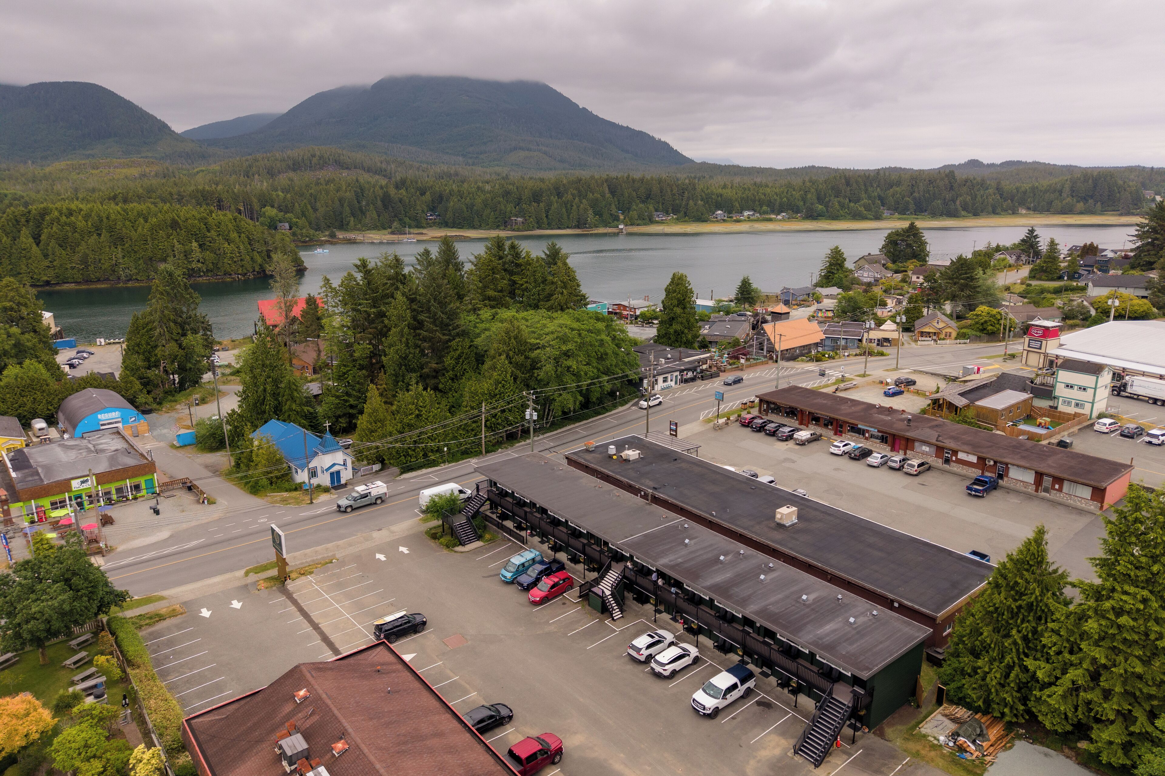 A commercial area with a parking lot, buildings, and a lake with mountains in the background.