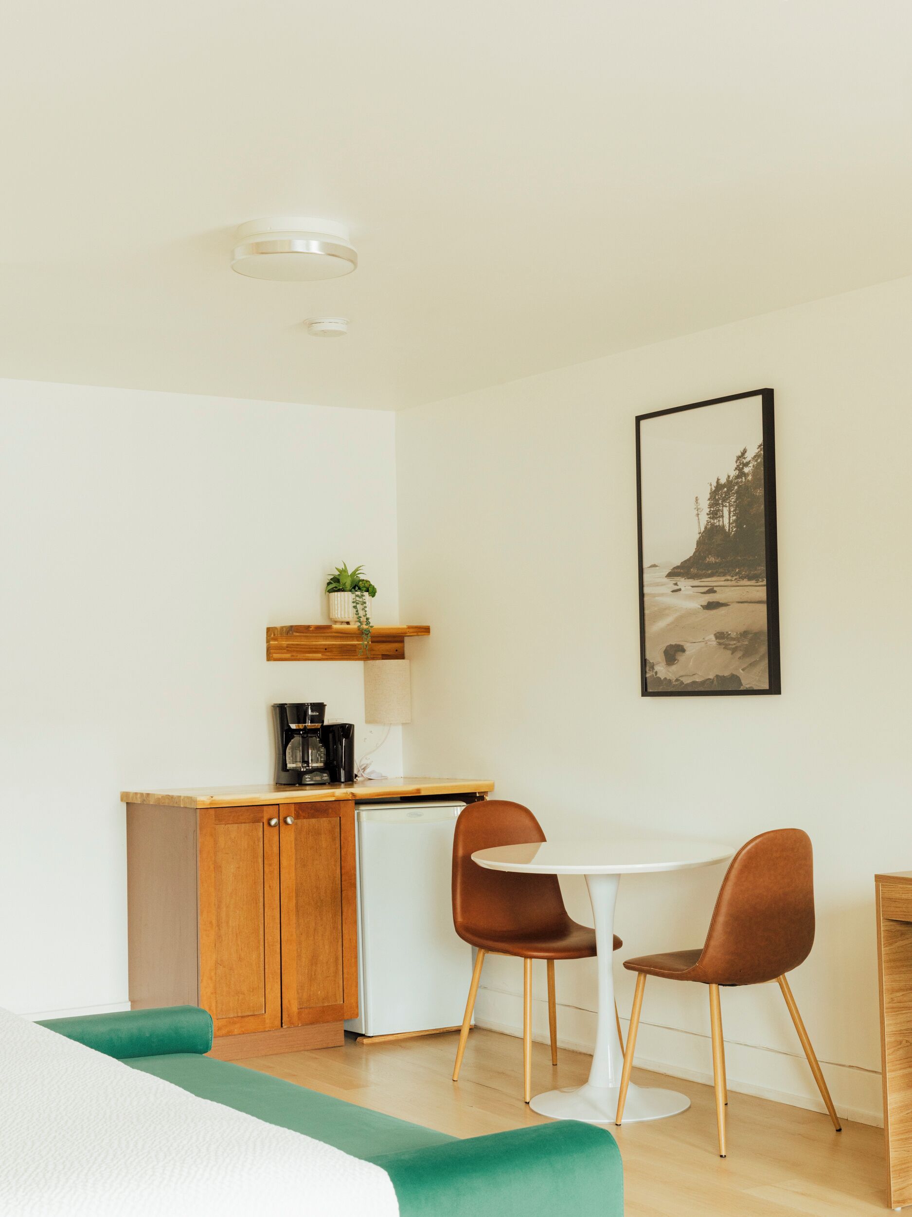 Una cocina compacta con cafetera, un refrigerador pequeño y un estante de madera con una planta. Hay una mesa blanca con dos sillas marrones y un cojín verde. En la pared cuelga un cuadro con una escena de playa.