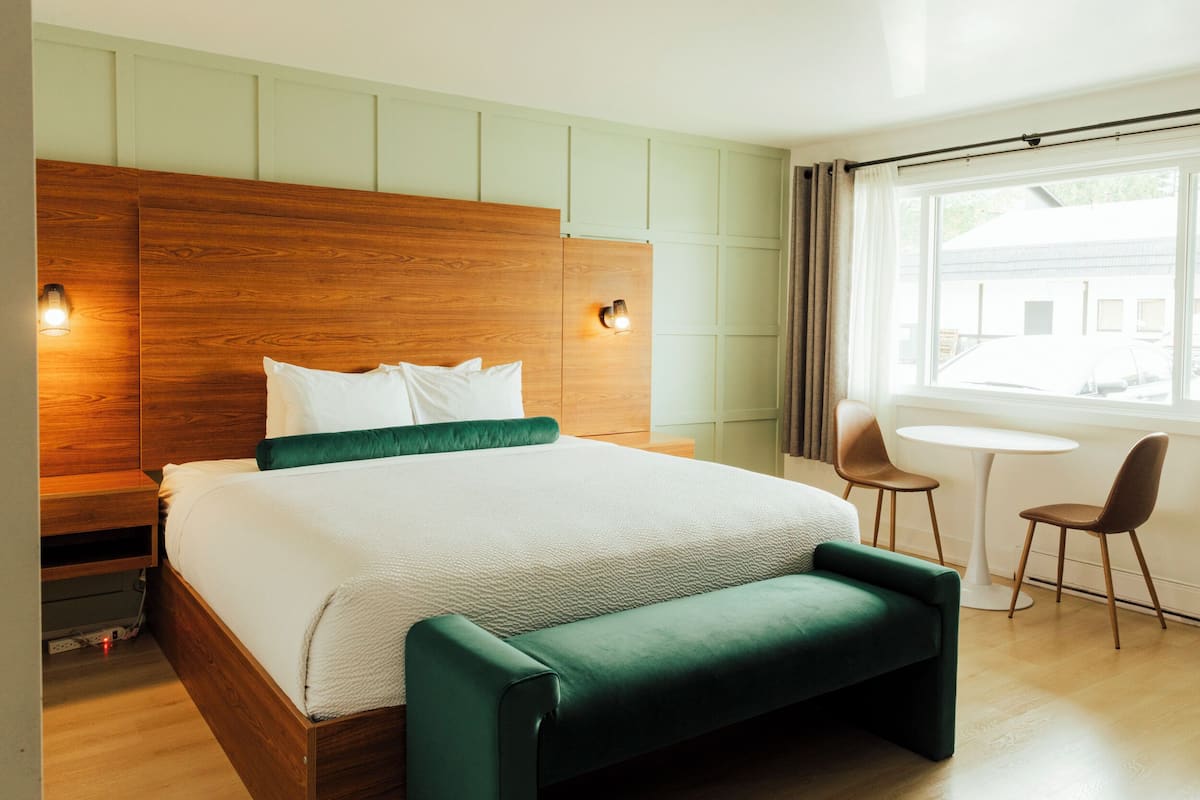 Ein modernes Hotelzimmer mit einem Holzkopfteil, einem großen Bett mit weißer Bettwäsche, einer gepolsterten grünen Bank, zwei Wandleuchten zum Lesen und einem kleinen Tisch mit zwei Stühlen.