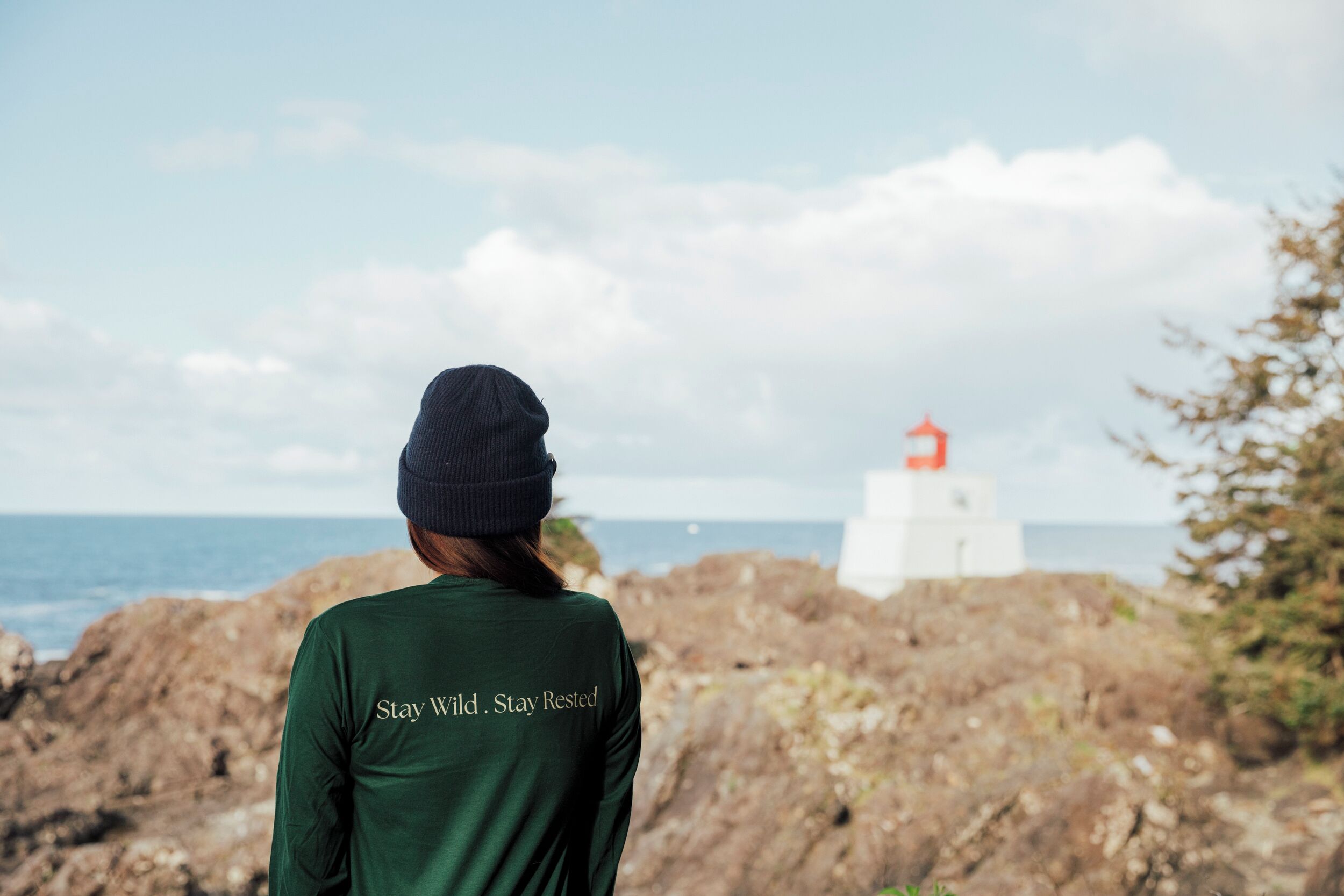 Eine Person mit grünem T-Shirt mit dem Aufdruck 'Stay Wild. Stay Rested.' steht an einer felsigen Küste, im Hintergrund ist ein Leuchtturm zu sehen.