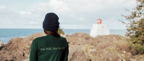 Eine Person mit grünem T-Shirt mit dem Aufdruck 'Stay Wild. Stay Rested.' steht an einer felsigen Küste, im Hintergrund ist ein Leuchtturm zu sehen.