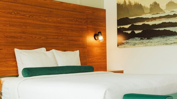 A bedroom with a wooden headboard, white bedding, a green throw pillow, a green bench, and a painting of a landscape on the wall.