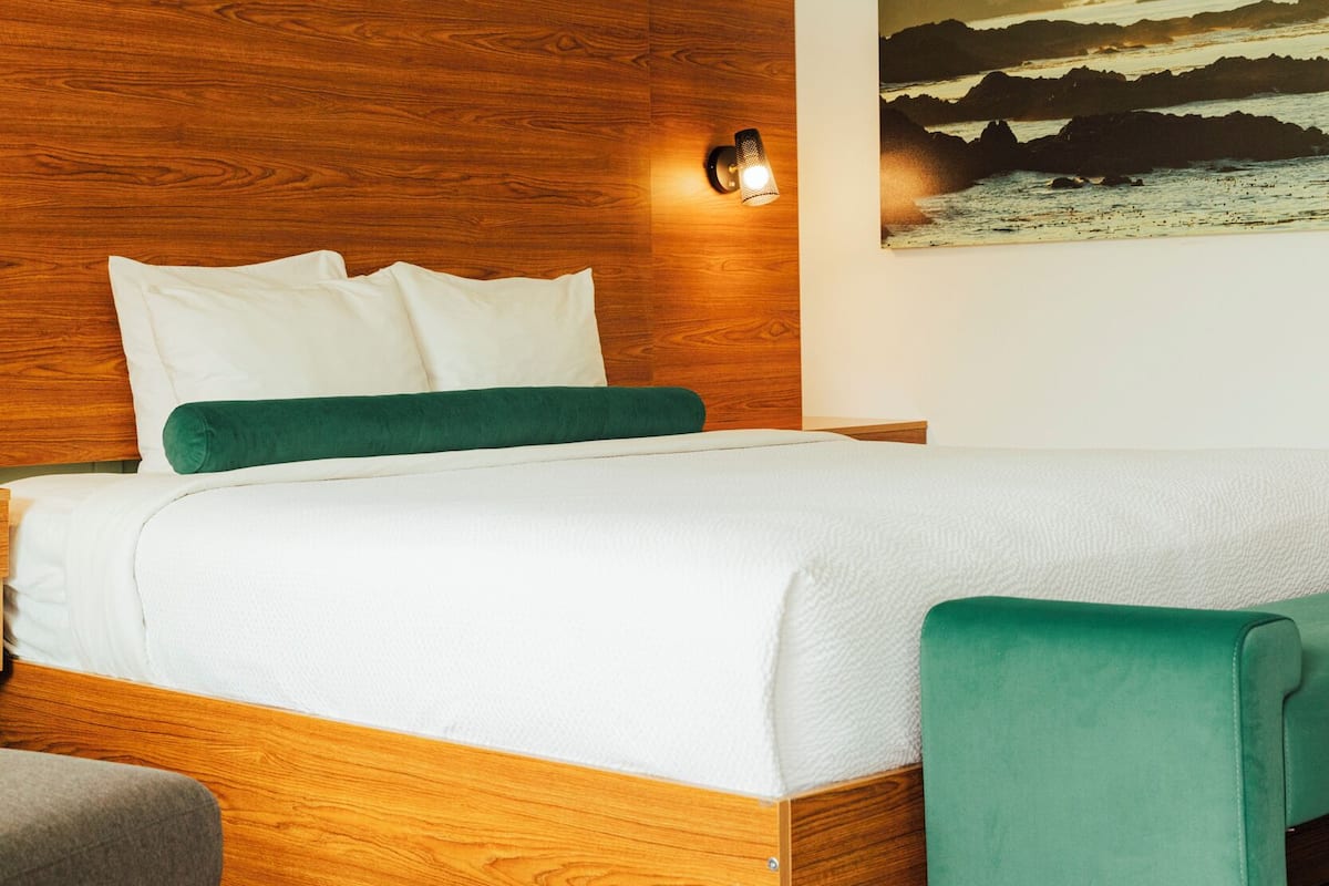 Ein Schlafzimmer mit einem Holz-Kopfteil, weißer Bettwäsche, einem grünen Zierkissen, einer grünen Bank und einem Landschaftsbild an der Wand.