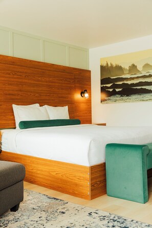 A bedroom with a wooden headboard, white bedding, a green throw pillow, a green bench, and a painting of a landscape on the wall. - Ucluelet Wildwood Inn (Ucluelet)