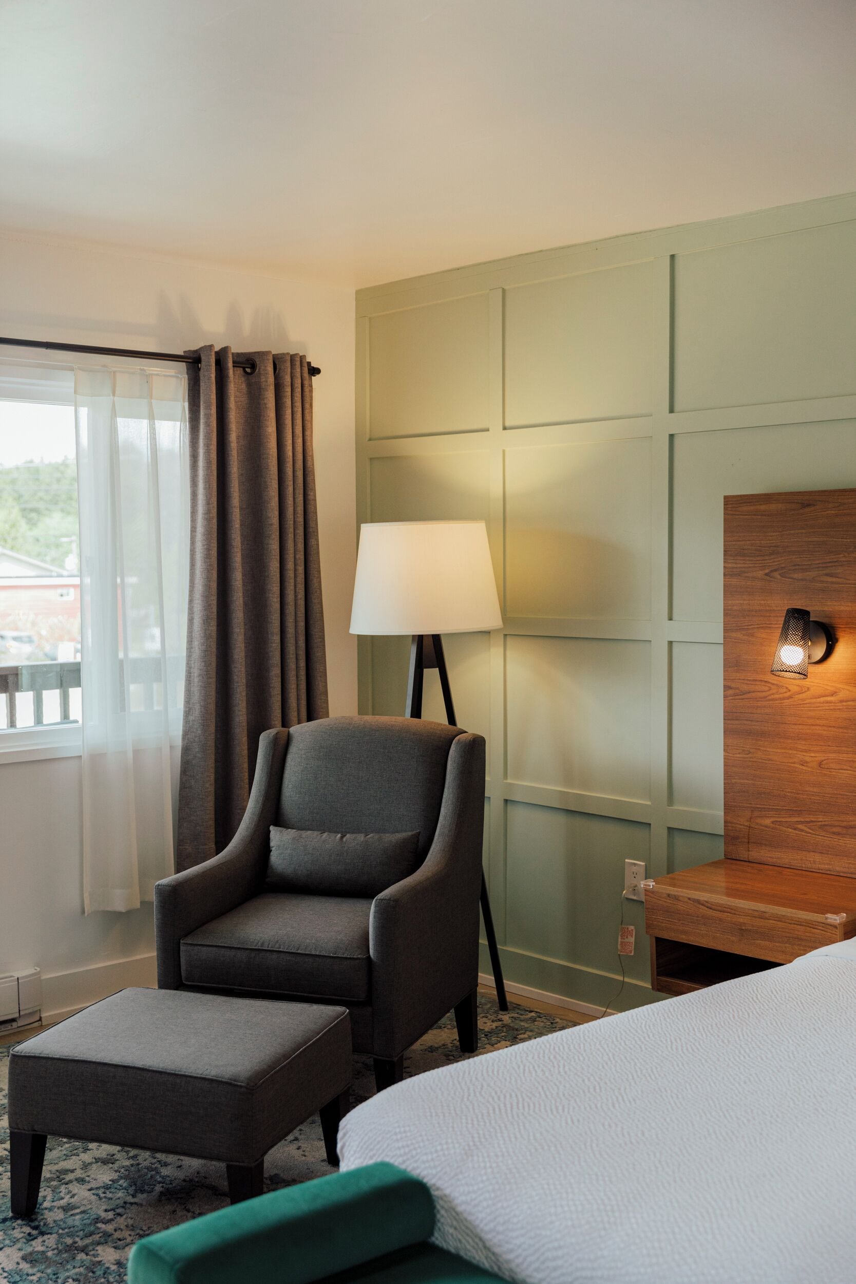 Camera d'albergo con un letto, una poltrona grigia, un poggiapiedi e una lampada, con una parete decorativa in legno.
