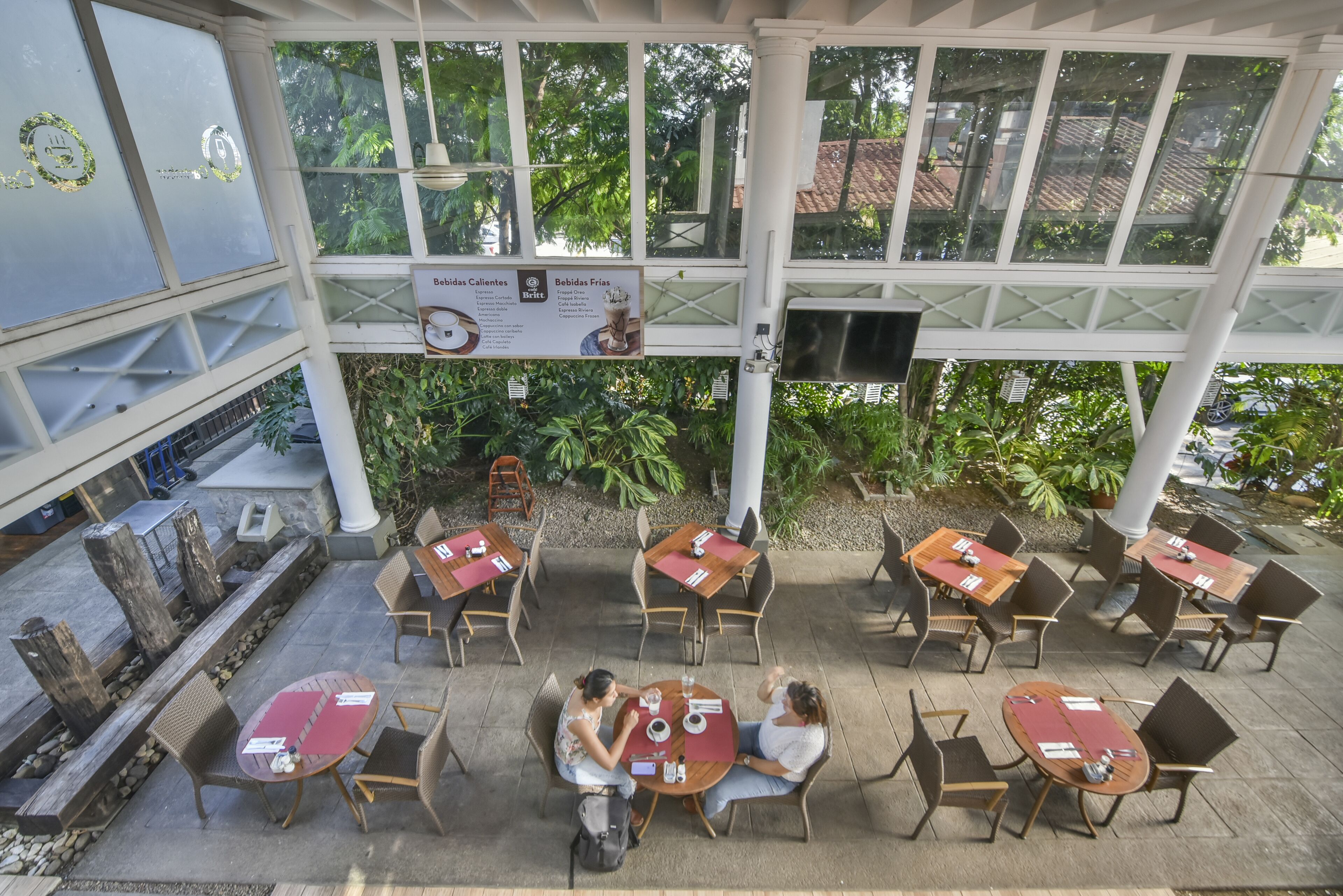 An outdoor cafe with tables and chairs, surrounded by greenery and a covered area with a TV screen.