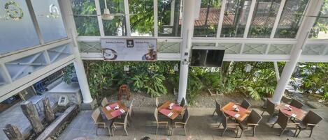 An outdoor cafe with tables and chairs, surrounded by greenery and a covered area with a TV screen.