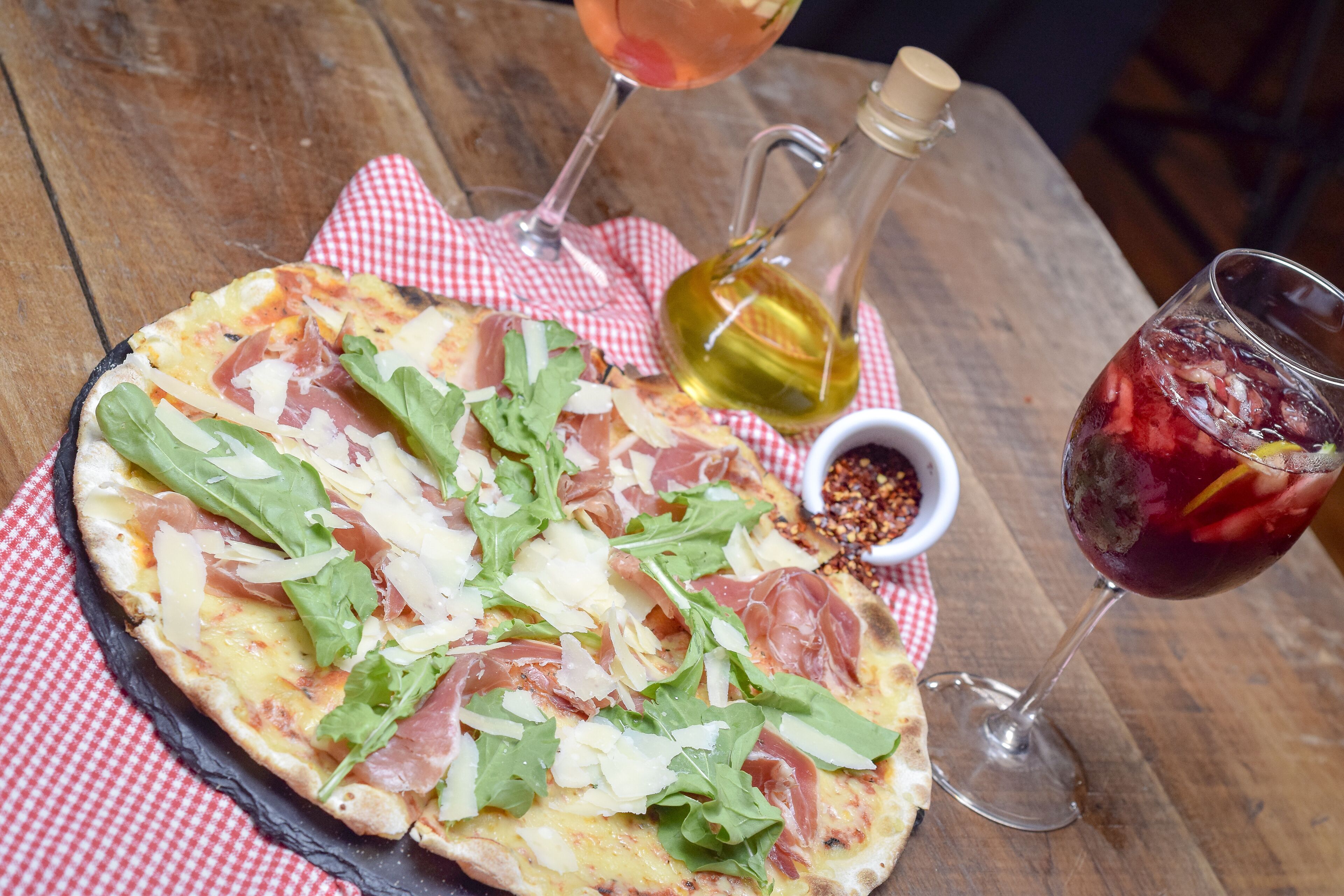 Pizza op een houten tafel met een rood-wit geruit servetje, een fles olijfolie en twee wijnglazen.