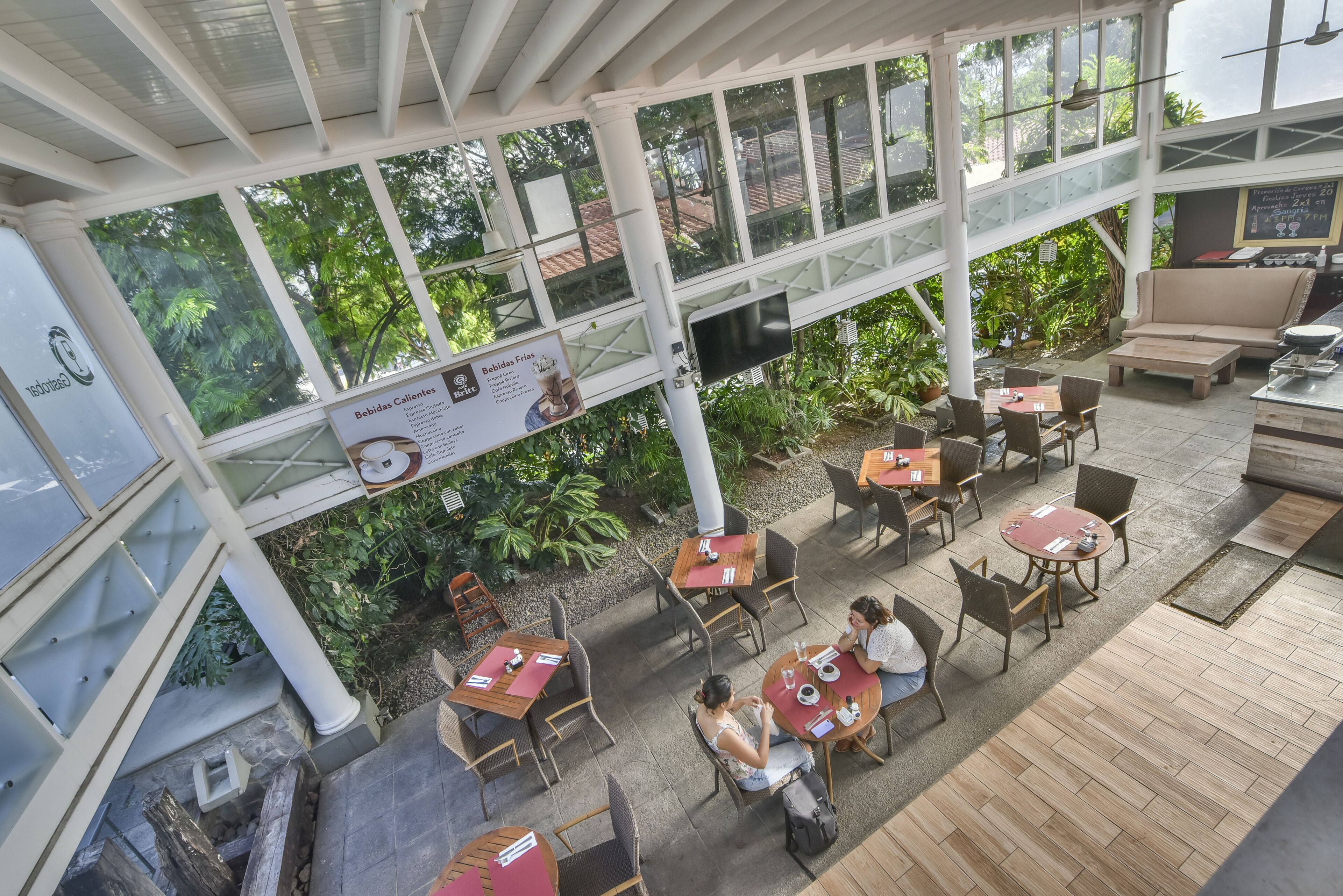 An outdoor café with tables and chairs, surrounded by greenery and large windows.