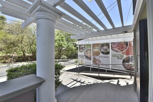 Un espacio al aire libre cubierto con una pérgola, un cartel publicitario de un restaurante y una carretera con vegetación de fondo.