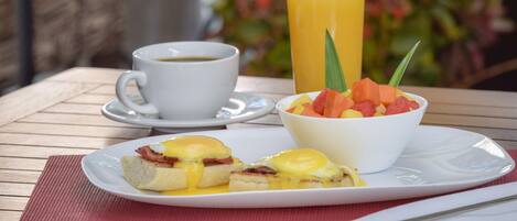 A breakfast setting with a plate of eggs Benedict, a glass of orange juice, and a bowl of fruit salad on a wooden table.