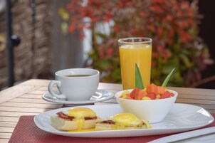 Un desayuno con un plato de huevos Benedict, un vaso de jugo de naranja y un bol de ensalada de frutas sobre una mesa de madera.