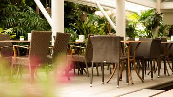 Een overdekt terras met rieten stoelen en tafels, omringd door weelderige beplanting.