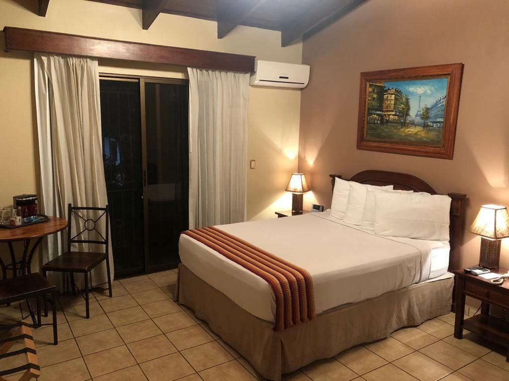 A hotel room with a bed, a desk, a chair, a table with a lamp, and a painting on the wall.