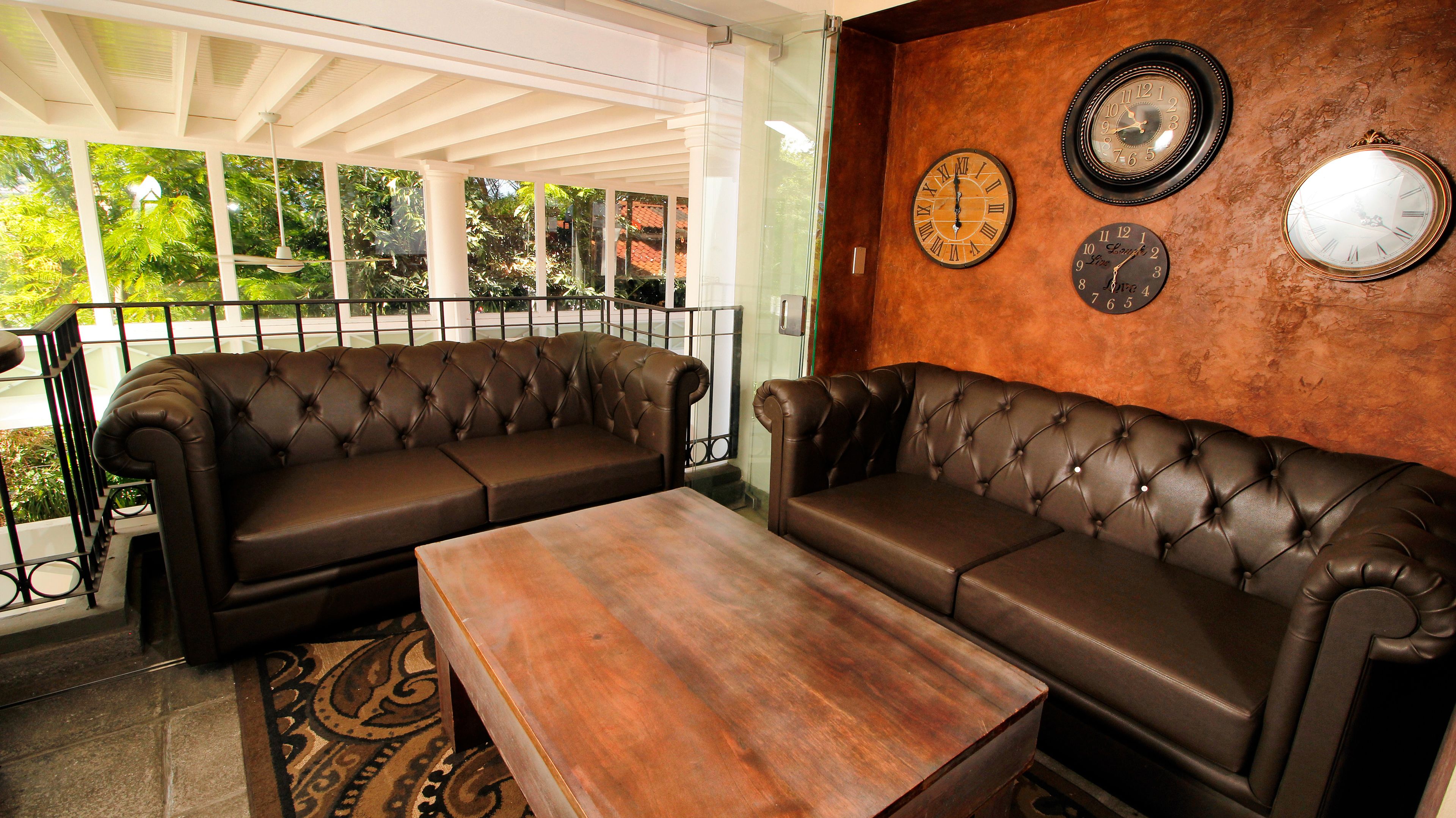 A cozy seating area with two brown leather sofas and a wooden coffee table, featuring a rustic wall with three clocks.