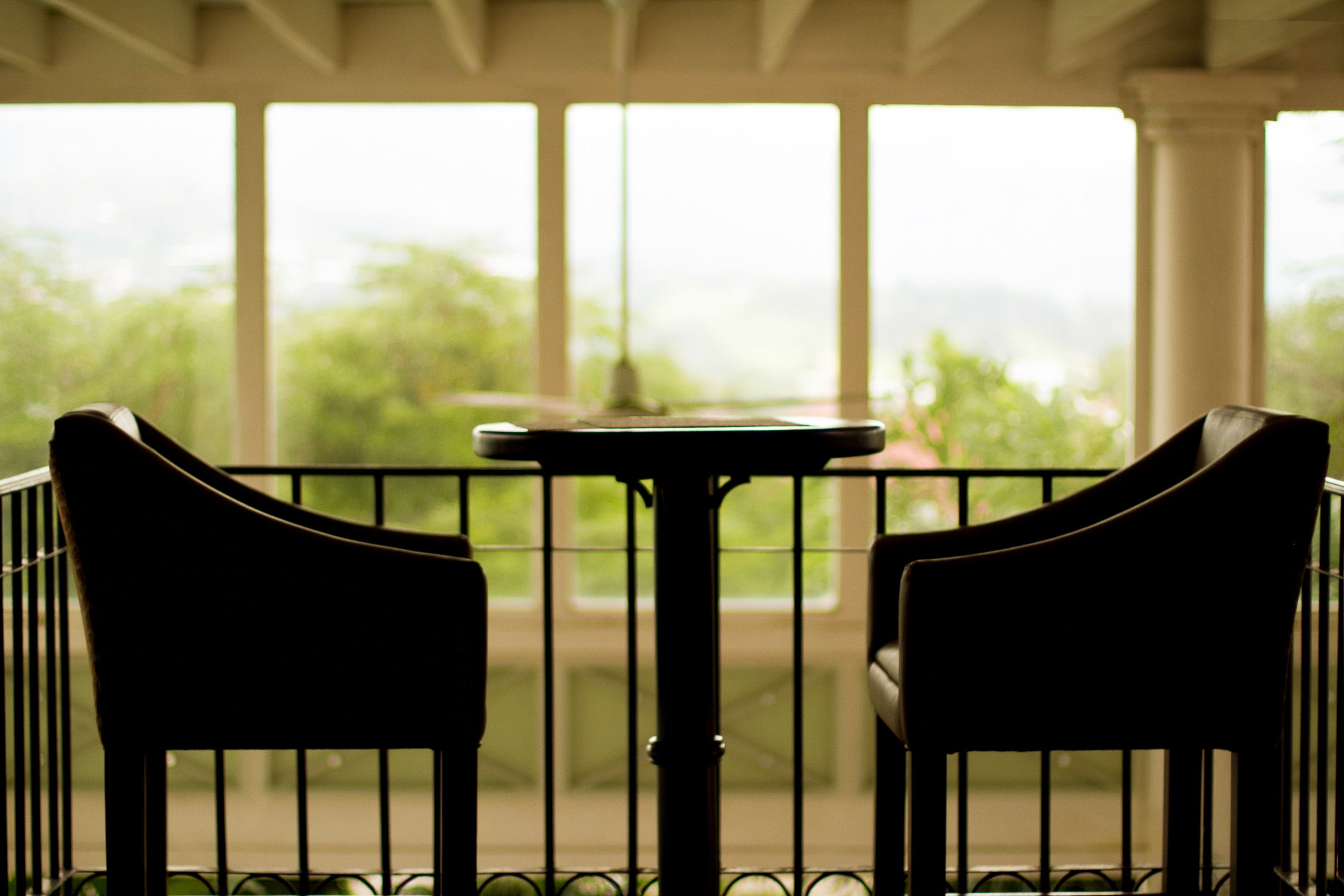 A covered outdoor seating area with two chairs and a table, featuring a view of greenery through large windows.
